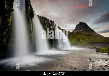 Atemberaubende Langzeitaufnahme der kaskadierenden Wasserfälle Islands mit dem berühmten Kirkjufell-Berg im Hintergrund unter einem pastellfarbenen Himmel. Stockfoto