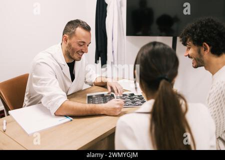 Ein fröhlicher männlicher Arzt erklärt Röntgenergebnisse einem jungen Paar in einer medizinischen Praxis, mit Schwerpunkt auf Patientenversorgung und Kommunikation. Stockfoto