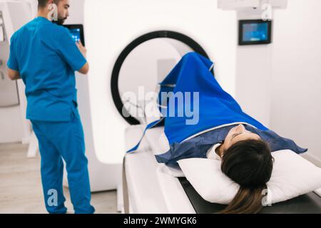 Ein Medizintechniker bedient einen CT-Scanner aufmerksam, während ein liegender Patient auf den Eingriff wartet, der mit einer blauen Decke bedeckt ist. Stockfoto