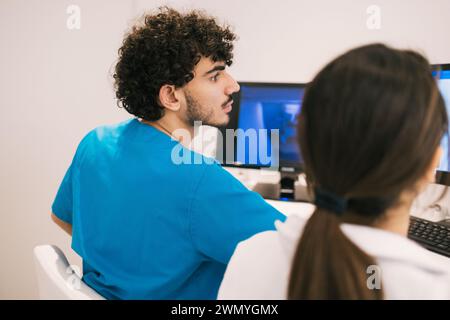 Ein männlicher medizinischer Mitarbeiter in blauem Scrubs bespricht Patientendaten mit einer Kollegin vor einem Computerbildschirm in einer klinischen Umgebung Stockfoto