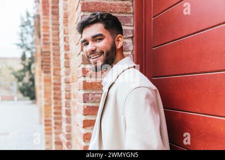 Charmanter Geschäftsmann in einem stilvollen Mantel, der sich an eine rote Ziegelwand lehnt und Selbstvertrauen und Leichtigkeit ausstrahlt Stockfoto