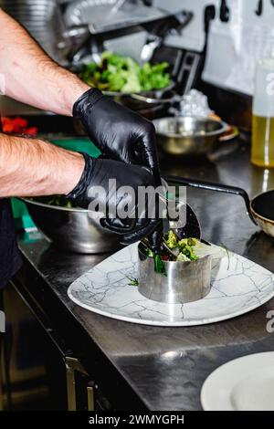 Nahaufnahme der Hände eines Küchenchefs mit schwarzen Handschuhen, die einen Gourmet-Salat in einem Metallring auf einem Marmorteller in einer professionellen Küche zart überziehen Stockfoto