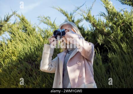 Frau in ihren 50ern fokussiert eine Kamera, um ein Foto zu machen, umgeben von Grün, zeigt einen Moment der Freizeit und des fotografischen Interesses Stockfoto