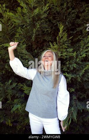 Überschwängliche reife Frau lacht und hebt ihre Faust vor einem Hintergrund von üppigem Grün Stockfoto