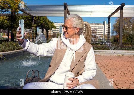 Reife Frau mit einer Kaffeetasse, die an einem sonnigen Tag ein Selfie an einem Brunnen macht, was einen aktiven urbanen Lebensstil widerspiegelt Stockfoto