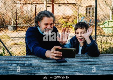 Ein Großvater und ein Enkel teilen einen freudigen Moment während eines Videogesprächs mit Familienmitgliedern draußen Stockfoto