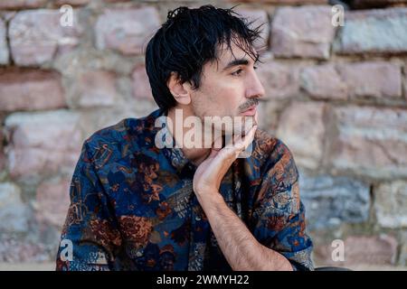 Ein Individuum erscheint tief in Gedanken, mit einem kontemplativen Ausdruck, Hand am Kinn, vor einer strukturierten Steinkulisse Stockfoto