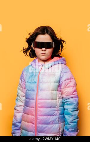 Ernstes junges Mädchen mit windgeblasenen Haaren, mit übergroßer futuristischer Brille und einer bunten Pufferjacke, vor gelbem Hintergrund Stockfoto