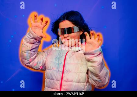 Kind in Pastelljacke mit erhobenen Händen, futuristische Brille auf blauem Hintergrund mit gelben Akzenten Stockfoto