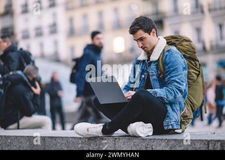 Der junge Mann arbeitet an seinem Laptop, während er auf der Treppe auf einem belebten Madrider Platz sitzt Stockfoto