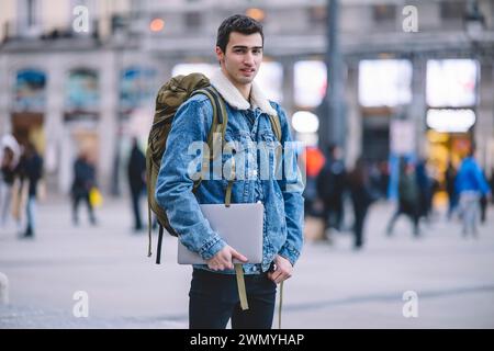 Ein selbstbewusster junger Mann, der einen Laptop hält, steht auf einem belebten Madrider Platz und blickt in die Kamera Stockfoto