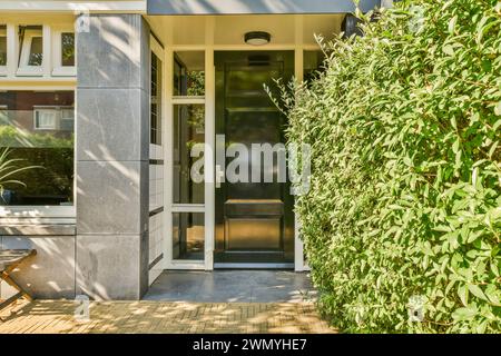 Ein moderner Hauseingang mit einer stilvollen schwarzen Tür, flankiert von großen Fenstern und einer leuchtend grünen Hecke. Stockfoto