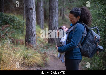 Eine lächelnde Mutter genießt eine Wanderung mit ihrem schlafenden Baby in einer vorderen Tragetasche, umgeben von einer ruhigen Waldlandschaft Stockfoto