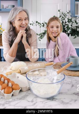 Schönes Mädchen, das Spaß mit ihrer Oma Frau in der Küche hat, die Teigprodukte zusammen zubereitet. Ältere Frauen geben Rezepte an jüngere Frauengattungen weiter Stockfoto
