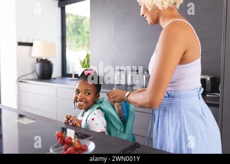 Eine junge birassische Mutter hilft einer afroamerikanischen Tochter mit ihrem Schulrucksack in einer Küche Stockfoto