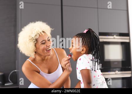 Junge birassische Mutter mit blonden lockigen Haaren lacht mit einer afroamerikanischen Tochter in einer Küche Stockfoto