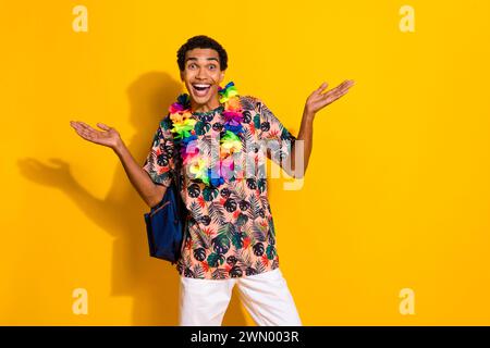 Foto von jungen funky positiven Kerl in exotischen Stil Blumen Halskette und Rucksack Touristen Reisende isoliert auf gelbem Hintergrund Stockfoto