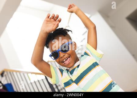 Der afroamerikanische Junge tanzt fröhlich, trägt eine blaue Sonnenbrille und ein gestreiftes Hemd Stockfoto