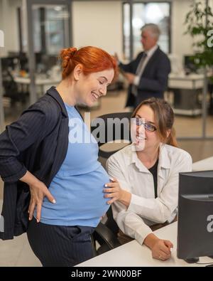Kaukasische Frau, die den Bauch eines schwangeren Kollegen berührt, während sie im Büro spricht. Stockfoto