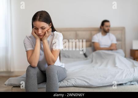 Besorgte Frau, die auf dem Bett sitzt, mit einem Mann im Hintergrund Stockfoto