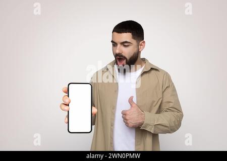 Ein überraschter junger Mann in einem beigefarbenen Hemd zeigt einen leeren Smartphone-Bildschirm mit der rechten Hand, während er mit den Daumen nach oben zeigt Stockfoto