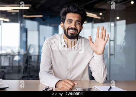 Online-Geschäftstreffen und -Schulungen. Ein junger indischer Geschäftsmann sitzt am Schreibtisch im Büro, winkt der Kamera und lächelt. Stockfoto