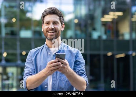 Nahaufnahme eines jungen lächelnden Mannes, der vor einem Bürozentrum auf der Straße steht, ein Telefon hält und in die Kamera schaut. Stockfoto
