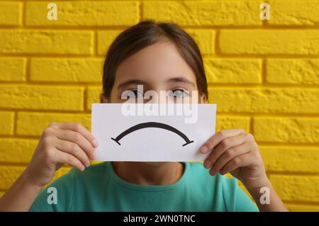 Trauriges Gesicht. Ein kleines Mädchen, das Gefühle zeigt, mit einer Karte gegen eine gelbe Ziegelwand Stockfoto