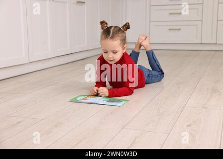 Niedliches kleines Mädchen liest Buch auf dem warmen Boden in der Küche, Platz für Text. Heizung Stockfoto