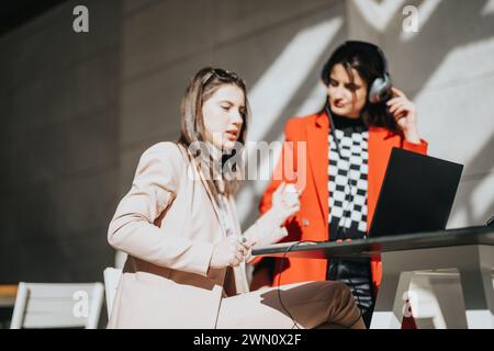 Zwei konzentrierte Geschäftsfrauen arbeiten an einem Projekt außerhalb zusammen, wobei eine einen Laptop und die andere über-Ohr-Kopfhörer anpasst. Stockfoto