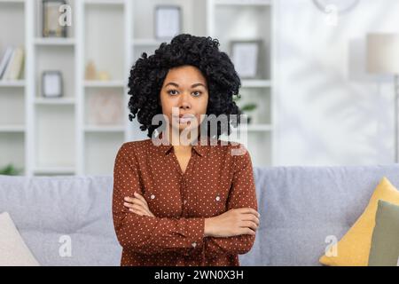 Porträt einer ernsten und selbstbewussten afroamerikanischen jungen Frau, die zu Hause auf der Couch sitzt und die Arme überkreuzt und in die Kamera blickt. Stockfoto
