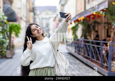 Lächelnde junge Indianerin, die auf der Straße der Stadt unterwegs ist und auf dem Handy Selfie macht, mit der Hand-zu-Handy-Kamera ein Siegeszeichen zeigt. Stockfoto