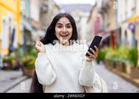 Porträt eines glücklichen und aufgeregten jungen indischen Mädchens in einem weißen Pullover, der mitten auf einer Stadtstraße steht und ein Telefon in der Hand hält, Erfolg und Nachrichten vor der Kamera genießt. Stockfoto