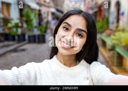 Porträt einer lächelnden jungen Indianerin in einem weißen Pullover, die ein Selfie auf einer Stadtstraße macht, online mit der Kamera spricht und ein Telefon hält. Stockfoto