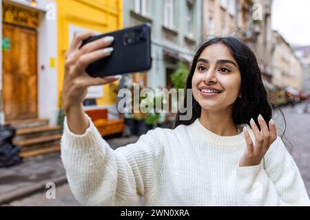 Nahaufnahme eines lächelnden jungen indischen Mädchens, das auf einer Stadtstraße läuft, ein Selfie macht und über einen Videoanruf telefoniert. Stockfoto