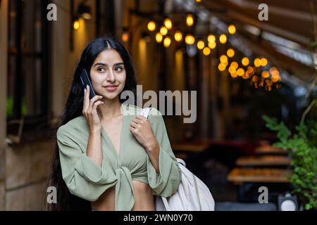 Eine junge indische Frau im grünen Anzug und mit Rucksack steht auf der Straße der abendlichen Stadt vor dem Hintergrund des Lichtes beim Telefonieren. Stockfoto