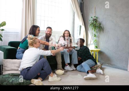 Eine Gruppe fröhlicher Freunde sitzt auf dem Boden und hebt ihre Gläser für einen Toast in einem lichtdurchfluteten Raum, in dem Wärme und Glück verkörpert werden Stockfoto