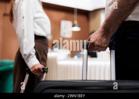 Aus nächster Nähe sehen wir, dass ein Paar mit Trolley-Taschen das Foyer eines schicken Hotels betritt. Detaillierte Aufnahme von älteren Personen, die Koffer greifen, als sie zum Registrierungsschalter gehen, um sich bei den Rezeptionisten einzuchecken. Stockfoto