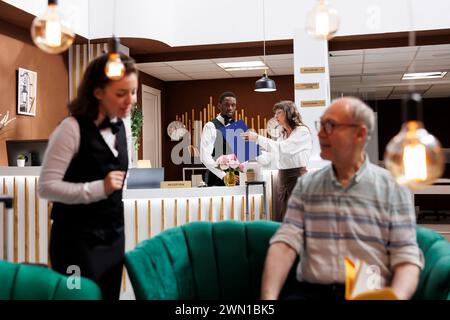 Die Seniorin meldet sich an der Hotelrezeption an, die auf einen angenehmen Urlaub begierig ist. Eine elterne Kundin füllt Reservierungsunterlagen aus, während der freundliche Concierge den Reisenden, der auf dem Sofa sitzt, rät. Stockfoto