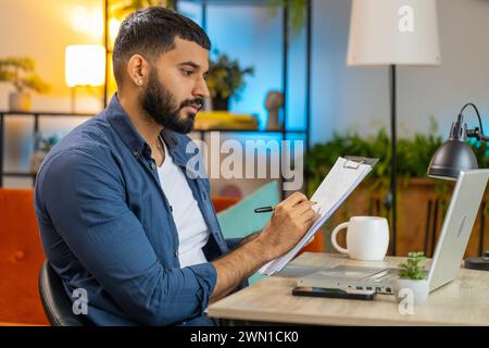 Indischer junger bärtiger Freiberufler macht Notizen in der Zwischenablage im Home Office, benutzt Laptop, liest Informationen und bereitet Geschäftsfinanzbericht vor. Der Typ, der Berichte analysiert, berechnet Steuern am Tisch. Stockfoto