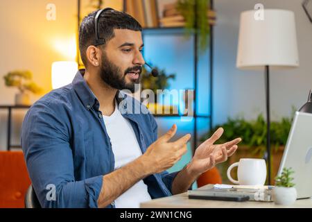 Indischer bärtiger Mann, der an einem Laptop arbeitet und ein Headset trägt freiberuflicher Mitarbeiter Callcenter, Support Service Operator Helpline mit angenehmer Kommunikation mit Kunden oder Kollegen im Home Office. Stockfoto