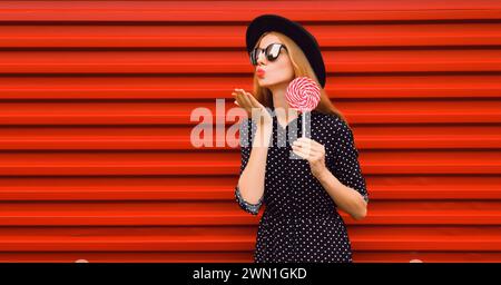 Sommerporträt einer glücklichen jungen Frau, die ihre Lippen bläst, die Luftküsse mit bunten Lutscher sendet, die einen schwarzen runden Hut auf rotem Hintergrund trägt Stockfoto