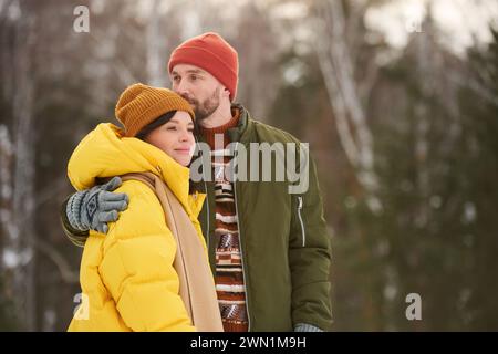 Mittleres Porträt eines verliebten jungen Mannes und einer verliebten Frau, die am Wintertag im Waldpark stehen, Mann, der den Kopf seiner Frau küsst Stockfoto