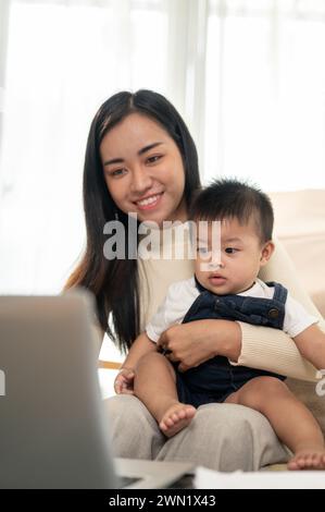 Eine schöne asiatische Geschäftsfrau, die Mutter, arbeitet von zu Hause aus, konzentriert sich auf ihren Laptop, während sie sich um ihren kleinen Sohn kümmert. Arbeiten Sie von zu Hause aus A Stockfoto