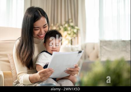 Eine glückliche asiatische Geschäftsfrau, die Mutter, konzentriert sich darauf, Dokumente zu lesen, von zu Hause aus zu arbeiten und sich um ihren süßen kleinen Sohn zu kümmern. Mamas Lebensrezept Stockfoto
