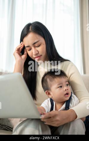 Eine unglückliche und beschäftigte asiatische Mutter hat Kopfschmerzen, während sie von zu Hause aus arbeitet und sich um ihren kleinen Sohn kümmert. Mamas Lebenskonzept Stockfoto