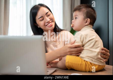 Eine glückliche asiatische Mutter spielt und füttert ihren kleinen Sohn mit Snacks, während sie in ihrem Heimbüro arbeitet. Arbeiten von zu Hause aus und die Lebenskonzepte von Mutter Stockfoto