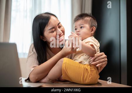 Eine glückliche asiatische Mutter spielt und füttert ihren kleinen Sohn mit Snacks, während sie in ihrem Heimbüro arbeitet. Arbeiten von zu Hause aus und die Lebenskonzepte von Mutter Stockfoto