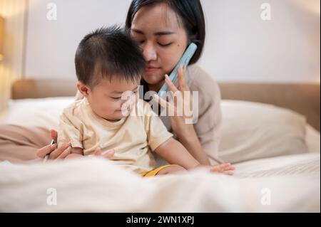 Ein süßer, unschuldiger asiatischer kleiner Junge spielt im Bett, während seine Mutter mit jemandem telefoniert. Kindheit und Mutterschaft Stockfoto