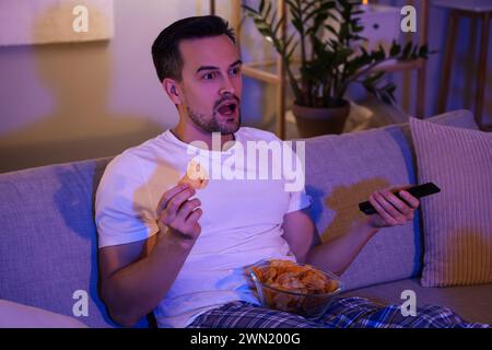 Schockierter Mann mit Kartoffelchips, der abends zu Hause fernsieht Stockfoto